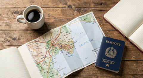 A folded paper map and a passport on a wooden table beside a small cup of coffee and an open notebook.