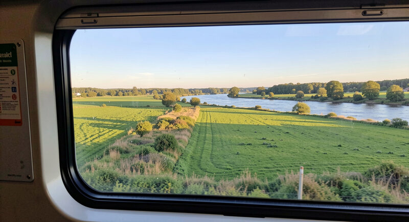 A train window view of green fields and a river passing under a clear morning sky.